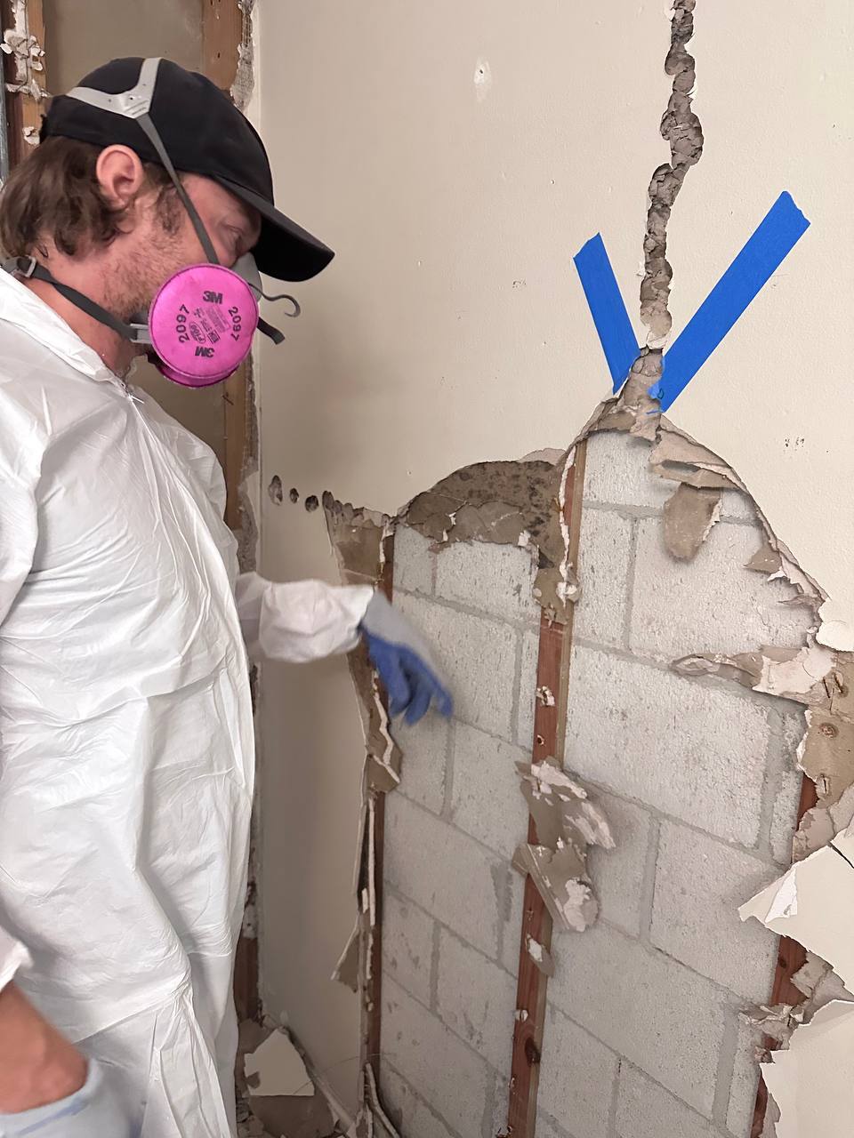 Technician in protective suit removing mold-damaged drywall from masonry wall